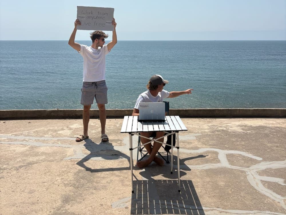 Einer von uns hält ein Schild hoch, der andere arbeitet am Laptop an einem Tisch direkt am Meer